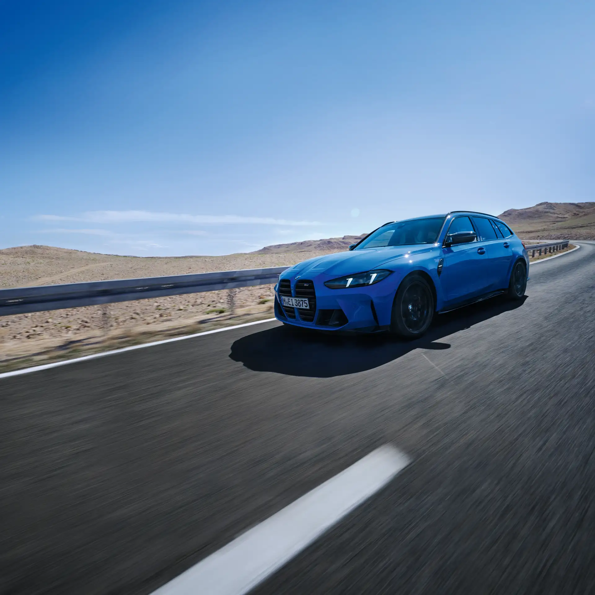 Blue BMW M3 Touring drives dynamically along a road through a stony desert landscape under a bright blue sky.