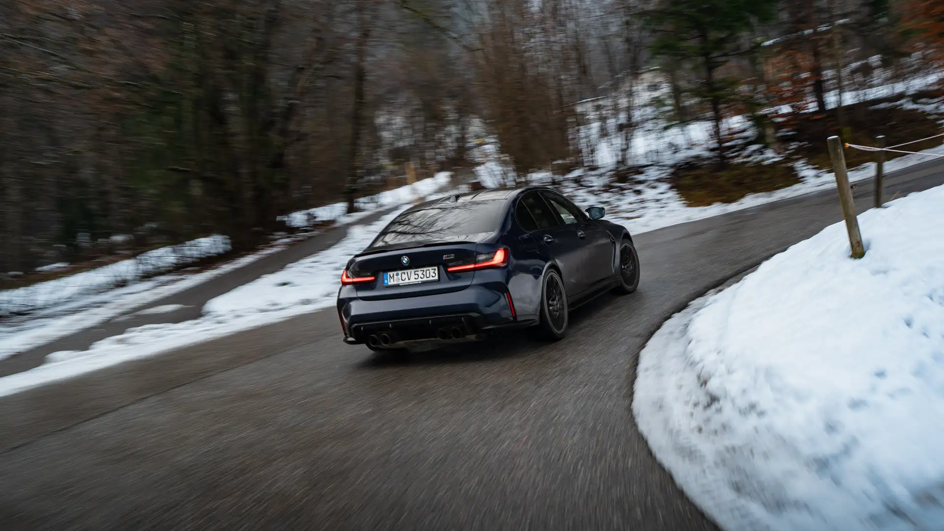 BMW M3 fährt schnell eine kurvige Serpentinenstraße am Tegernsee hinauf, auf nasser, asphaltierter Straße, mit dynamischer Kurvenfahrt und Bergkulisse.