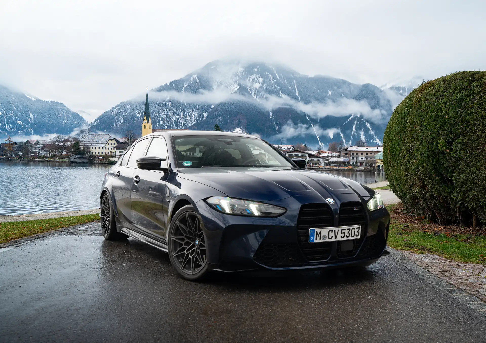 BMW M3 am Tegernsee im Winter, mit Blick auf den See, eine gelbe Kirche im Hintergrund und einen Berg, der oben von Nebel bedeckt ist.