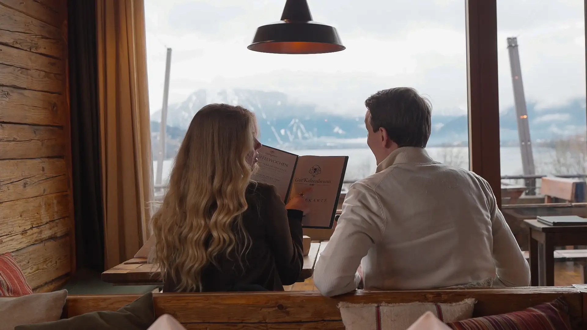Ein Mann und eine Frau sitzen im Gut Kaltenbrunn mit Blick auf den Tegernsee, schauen in die Speisekarte und überlegen, was sie bestellen möchten.