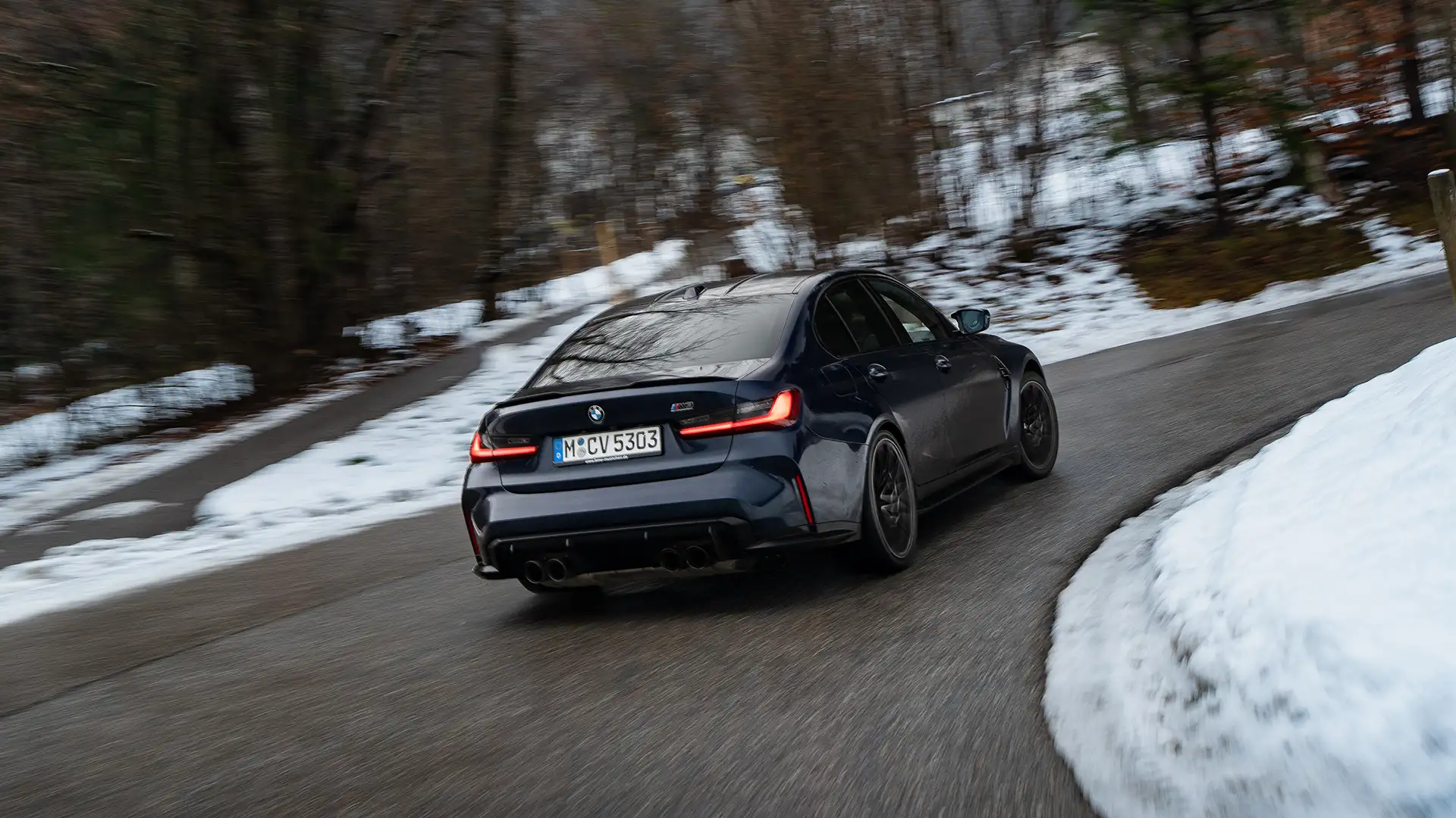 Dark blue BMW M3 driving uphill in a curve through a snowy landscape with trees in the background.