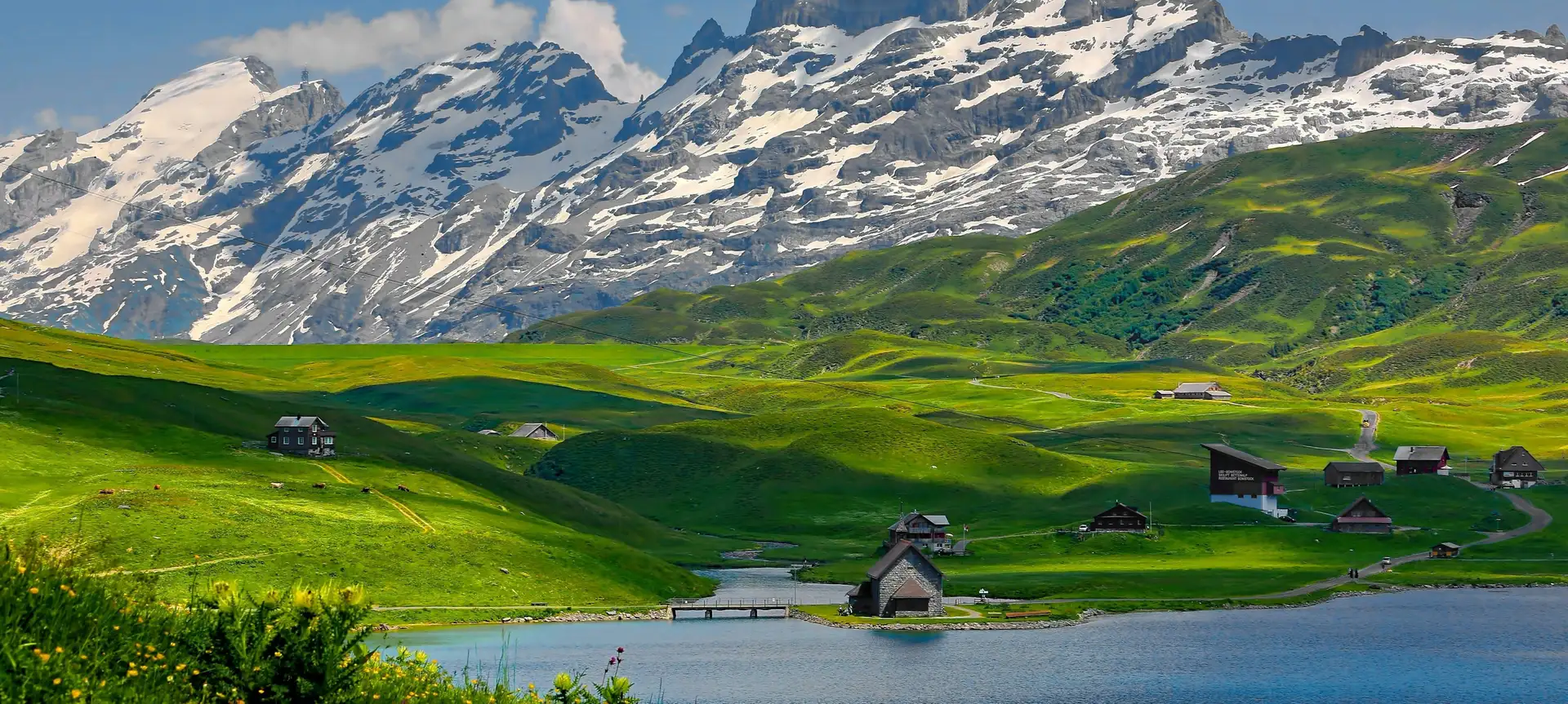 A mountain relief rises up behind green meadows and Lake Melchsee in Switzerland.